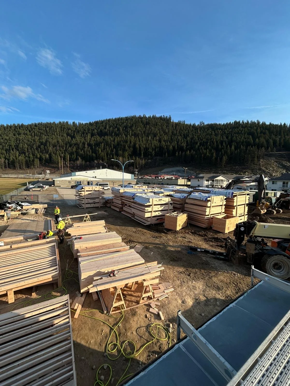 Construction site with workers, wooden materials, equipment, and machinery, with a backdrop of trees and a clear blue sky.