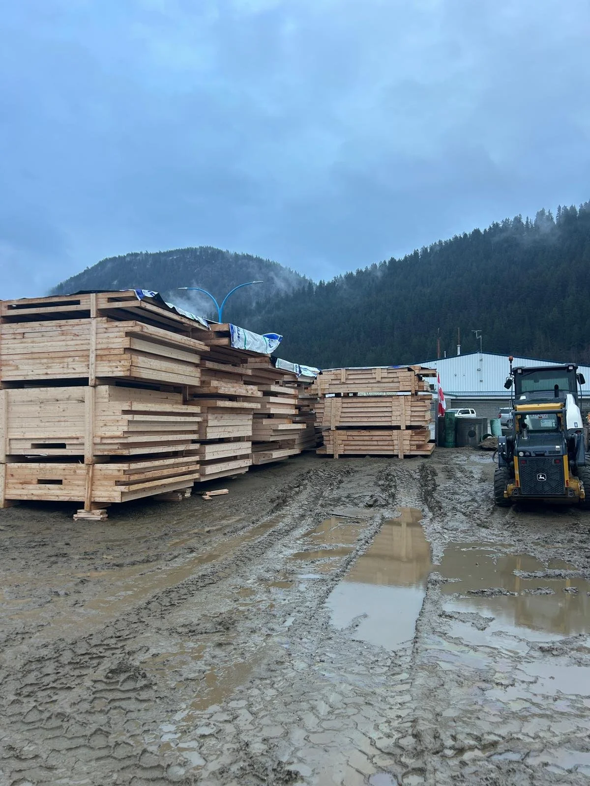 Stacks of wooden lumber stored outside at a construction site with muddy ground and puddles, a small construction vehicle, and a building with mountains in the background.