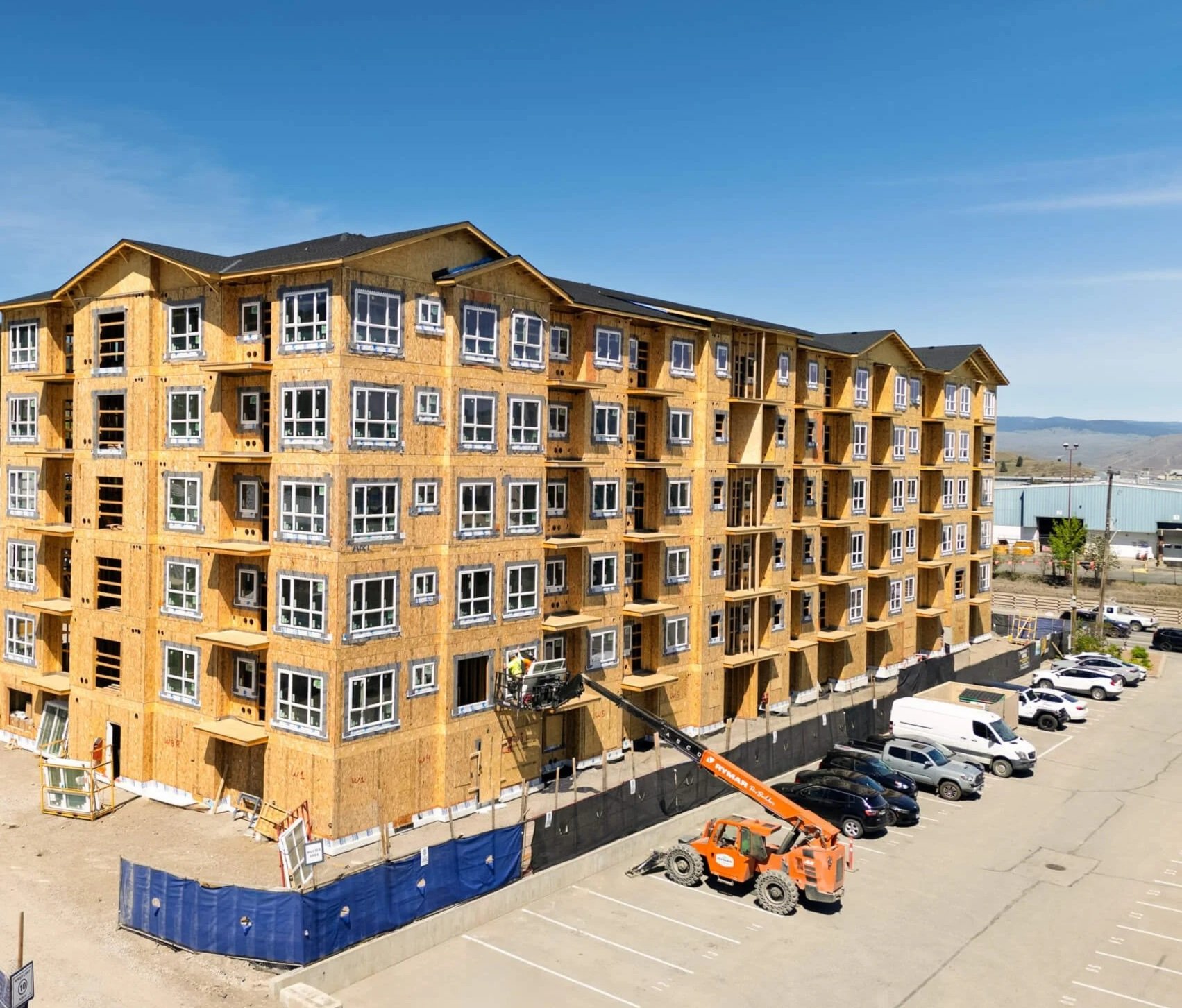 A multi-story building under construction with wooden framing and numerous windows, with a construction lift and several parked cars on a sunny day.