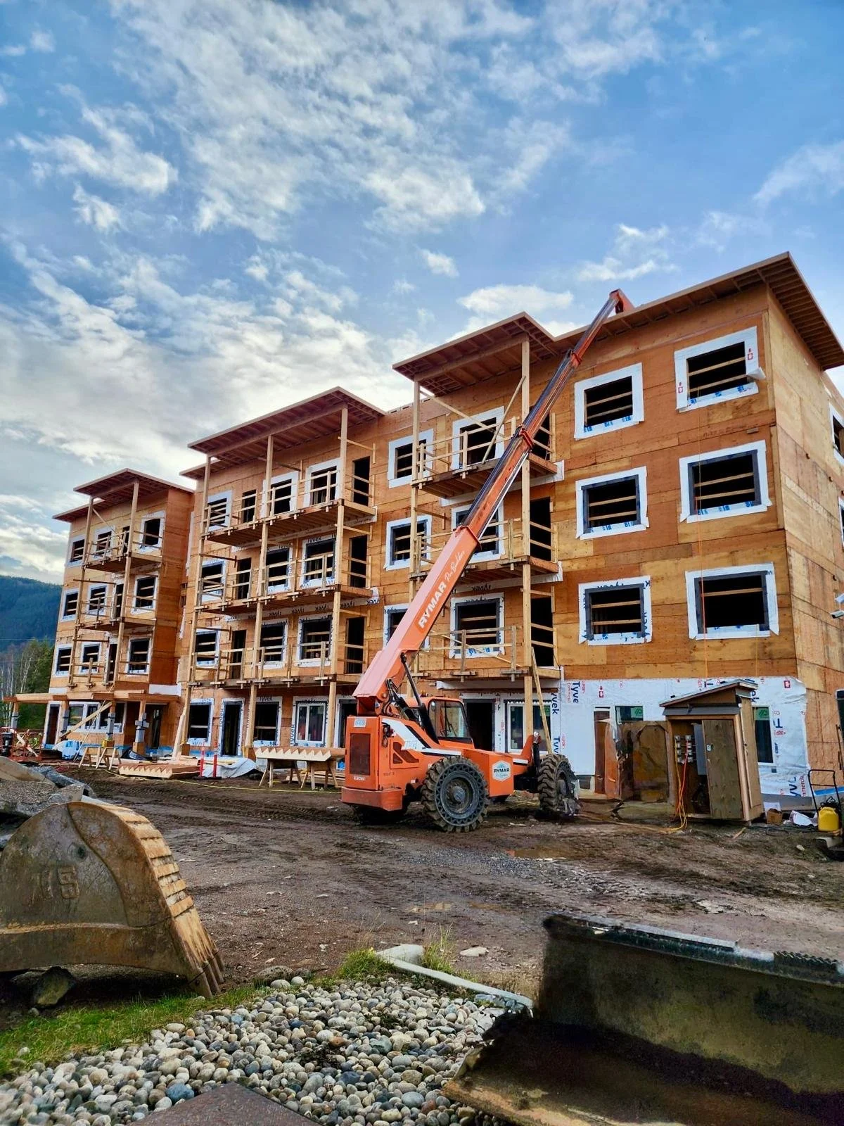 Construction site of a multi-story building with wooden framing, scaffolding, and a crane in use, under a partly cloudy sky.