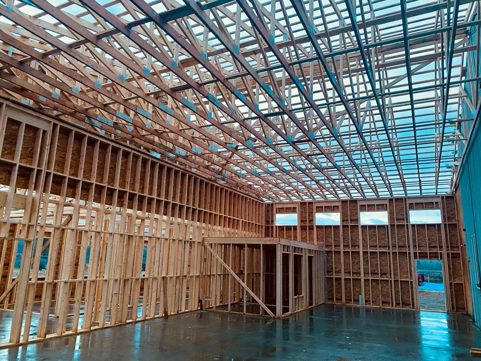 Interior view of a building under construction with exposed wooden framing on the walls and roof, and a concrete floor.