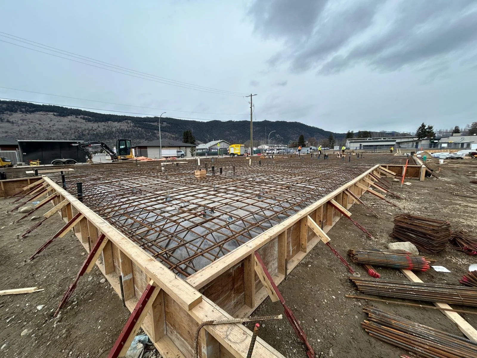 Construction site with a foundation frame and rebar, workers in the distance, overcast sky, mountains in the background.