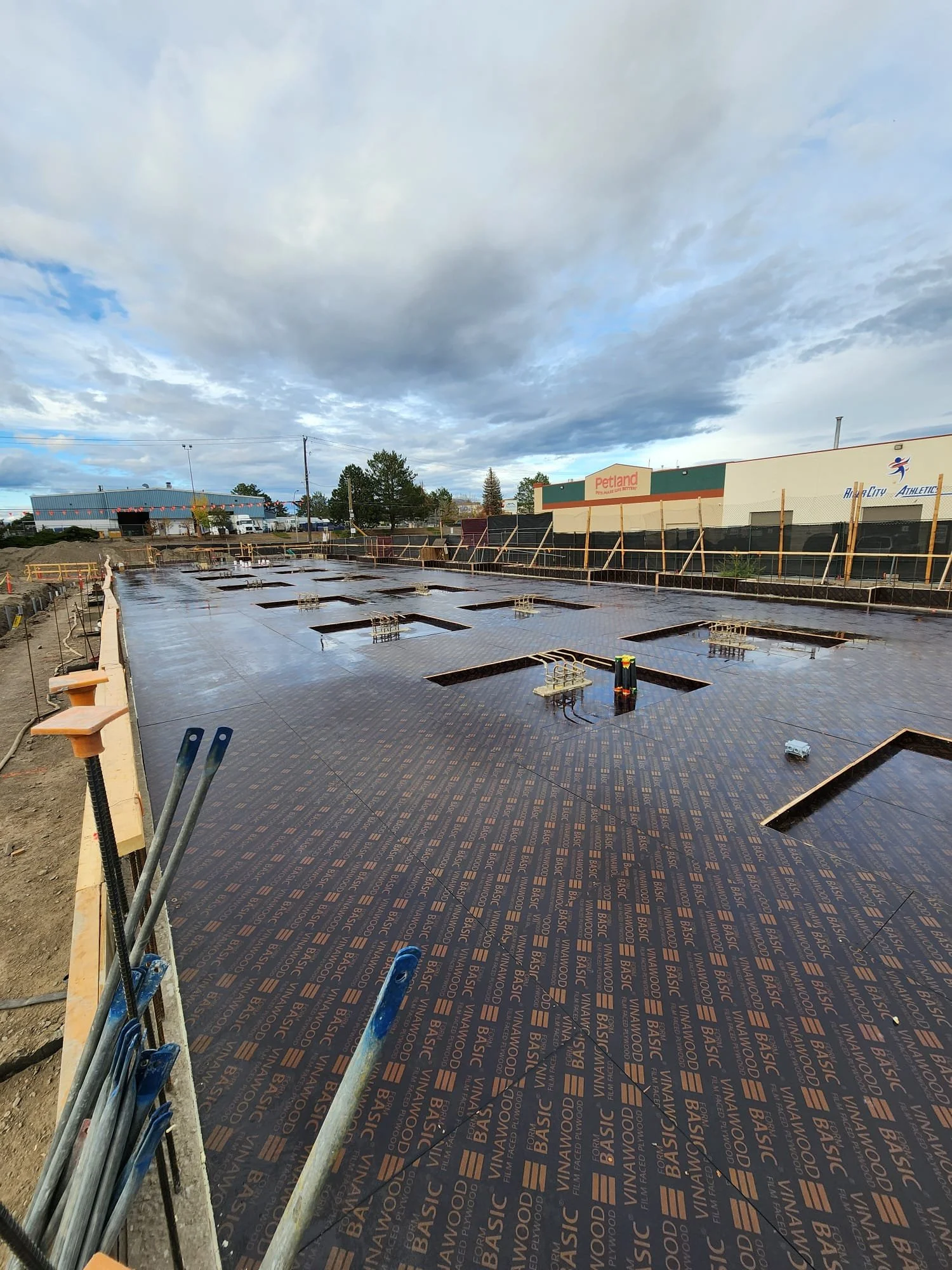 Construction site with a concrete foundation, metal rebar, and black plastic vapor barrier on the ground, under a cloudy sky.