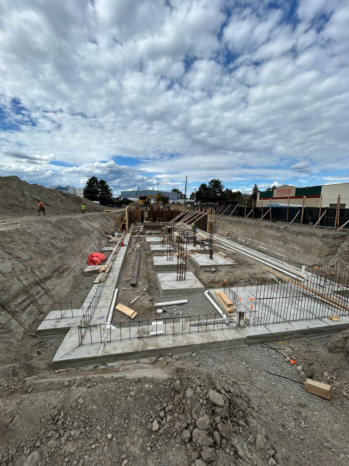 Construction site showing a foundation being built, with concrete slabs, rebar, and construction workers, under a partly cloudy sky.