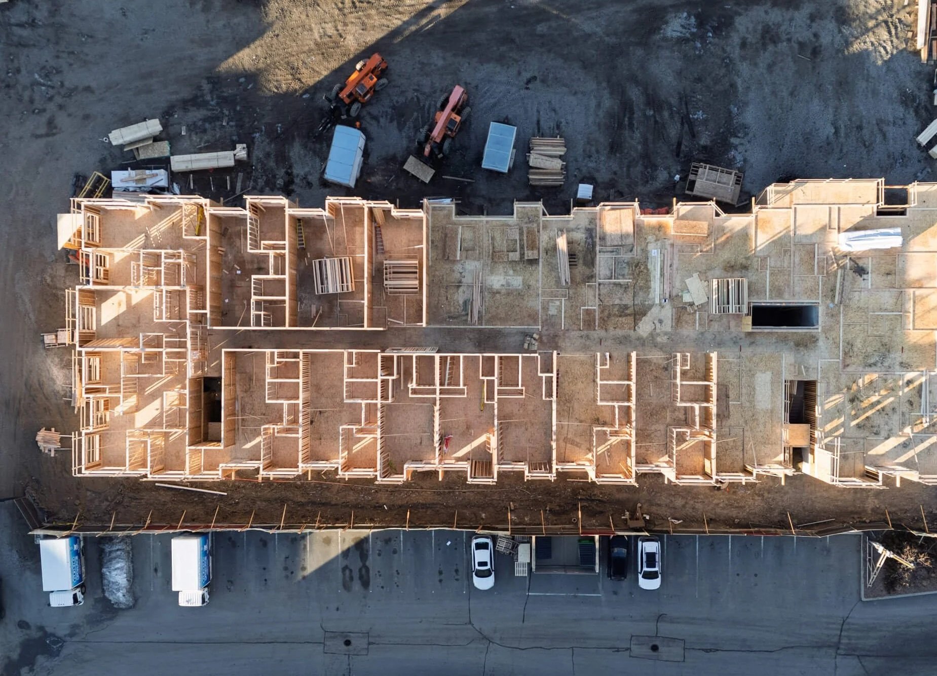 An aerial view of a multi-story building under construction, with wooden framing in place. Construction equipment and materials are visible at the site, and parked cars are seen along the street at the ground level.