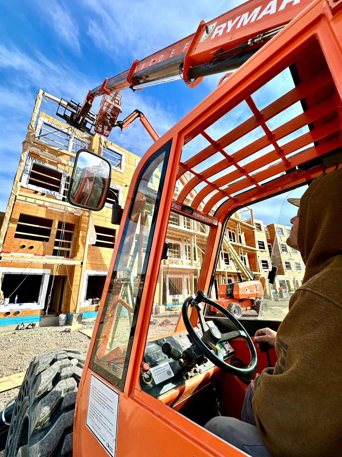 A construction worker operating an orange telescopic lift at a building site with wooden framing. The lift's controls are visible in the foreground, and scaffolding and building structures in the background under a blue sky.