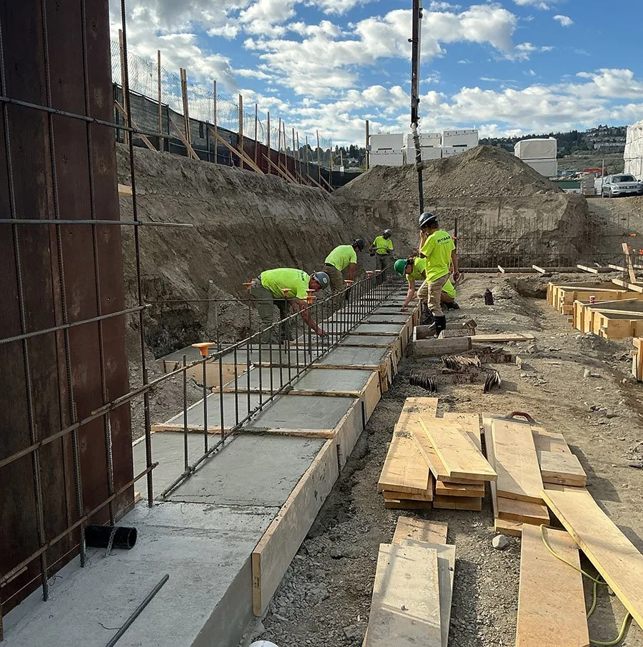 Construction workers pouring and setting concrete for a sidewalk or foundation on a construction site, with rebar and wooden framing in place, and dirt mounds in the background on a partly cloudy day.