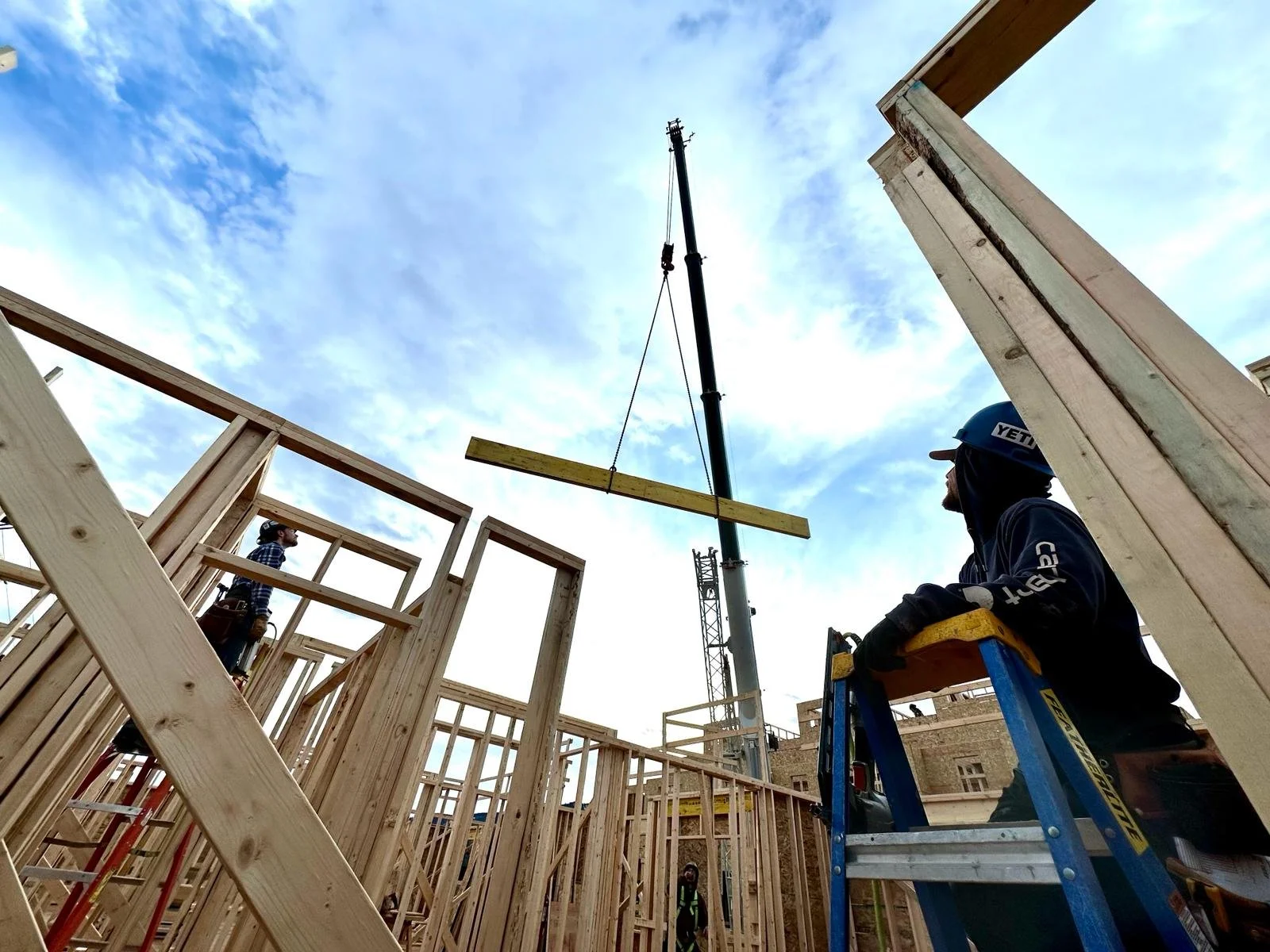 Construction workers building a wooden frame for a house with a crane lifting a beam into place under a cloudy sky.