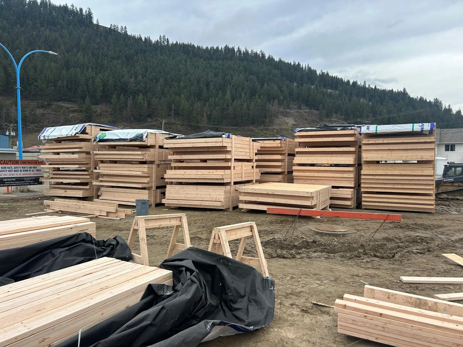 Construction site with prefabricated wall panels, some covered with plastic, in front of a mountain landscape, dirt ground, construction signs and equipment visible.