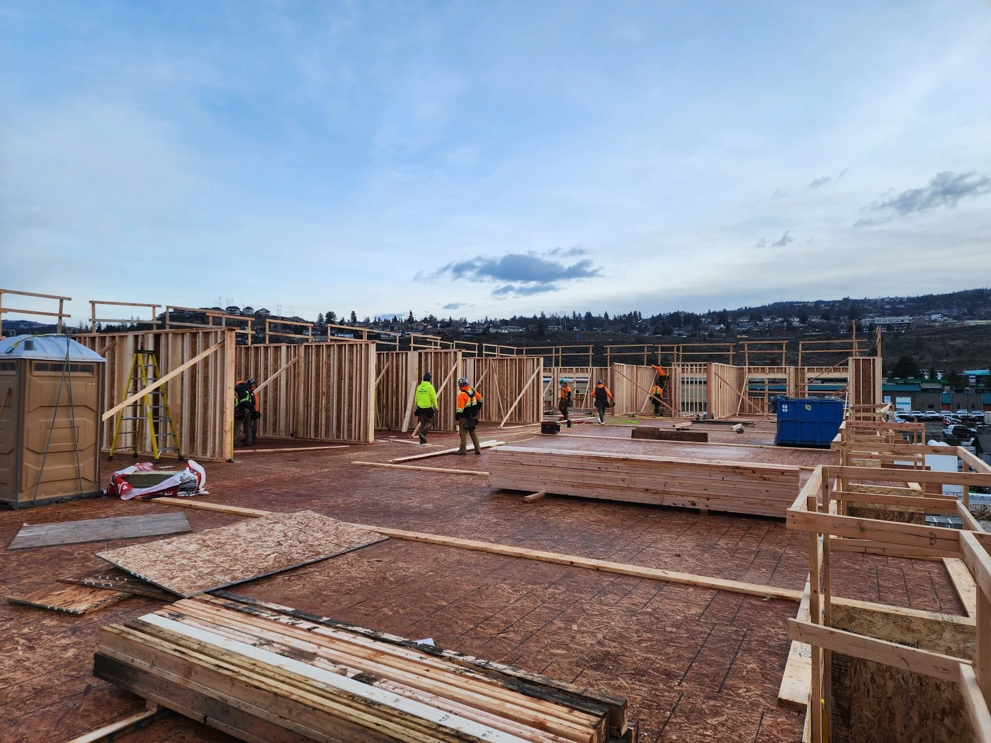 Construction workers building the wooden framework of a multi-story structure on a construction site with a cityscape in the background under a cloudy sky.