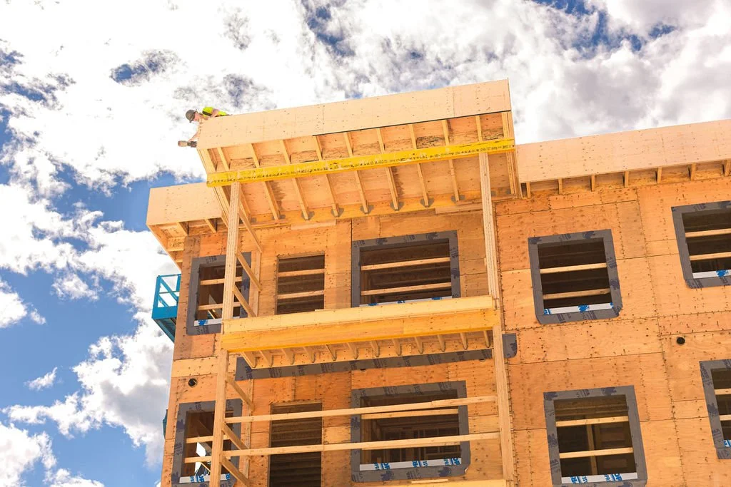A partially constructed multi-story building with wooden framing, open window spaces, and a blue sky with clouds in the background.