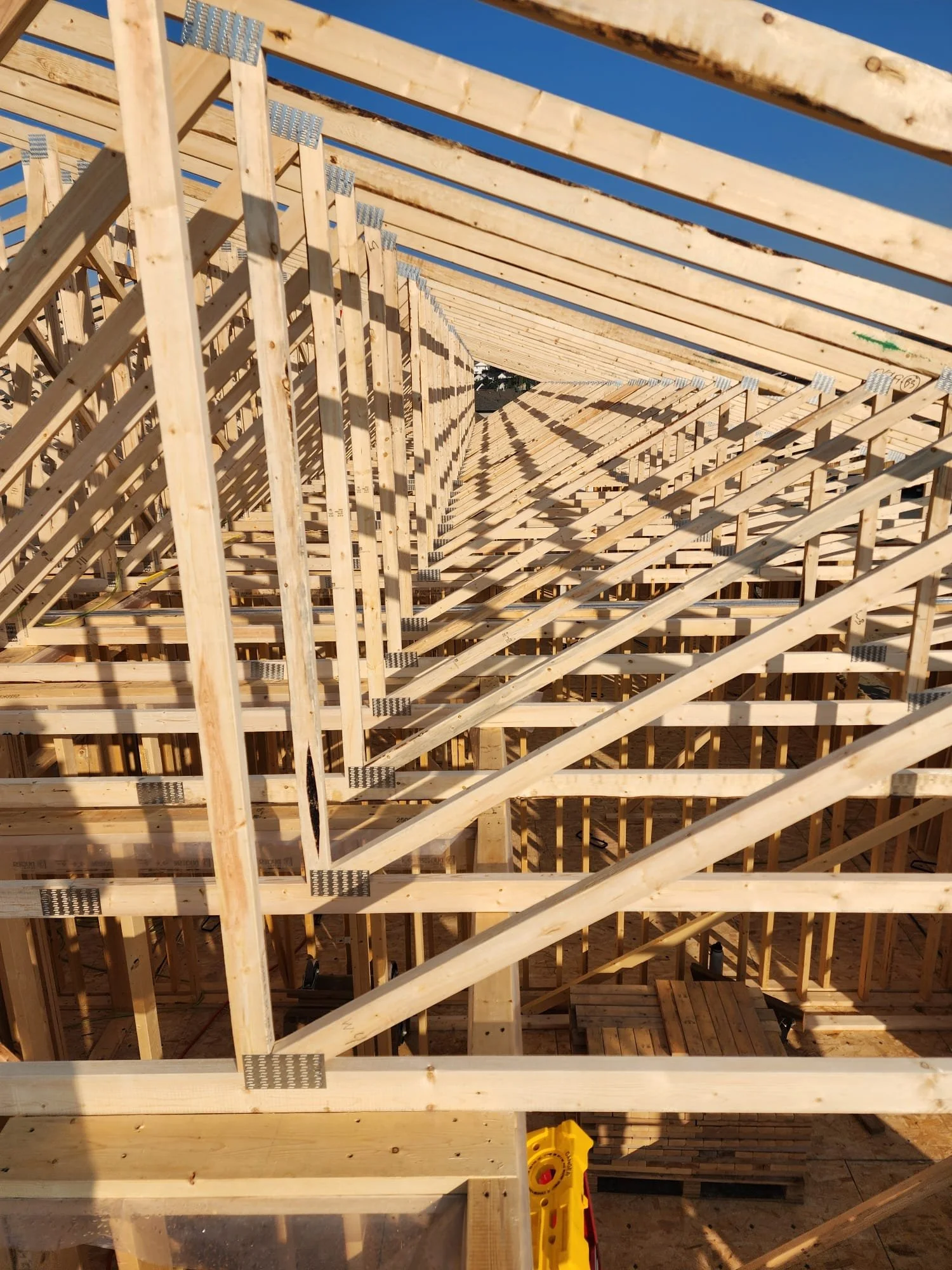 A wooden building framework under construction with visible roof rafters and support beams. Clear blue sky overhead and construction materials on the ground.