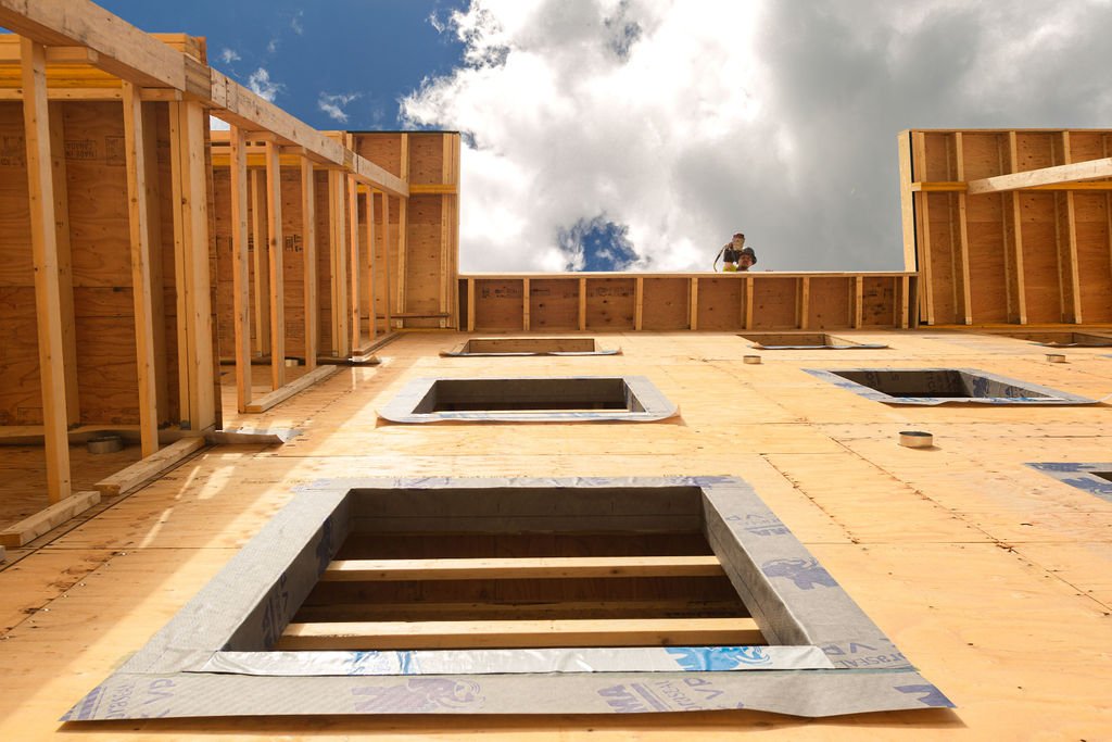 A building under construction with wooden framing and window openings, viewed from the ground looking up towards a partly cloudy sky.