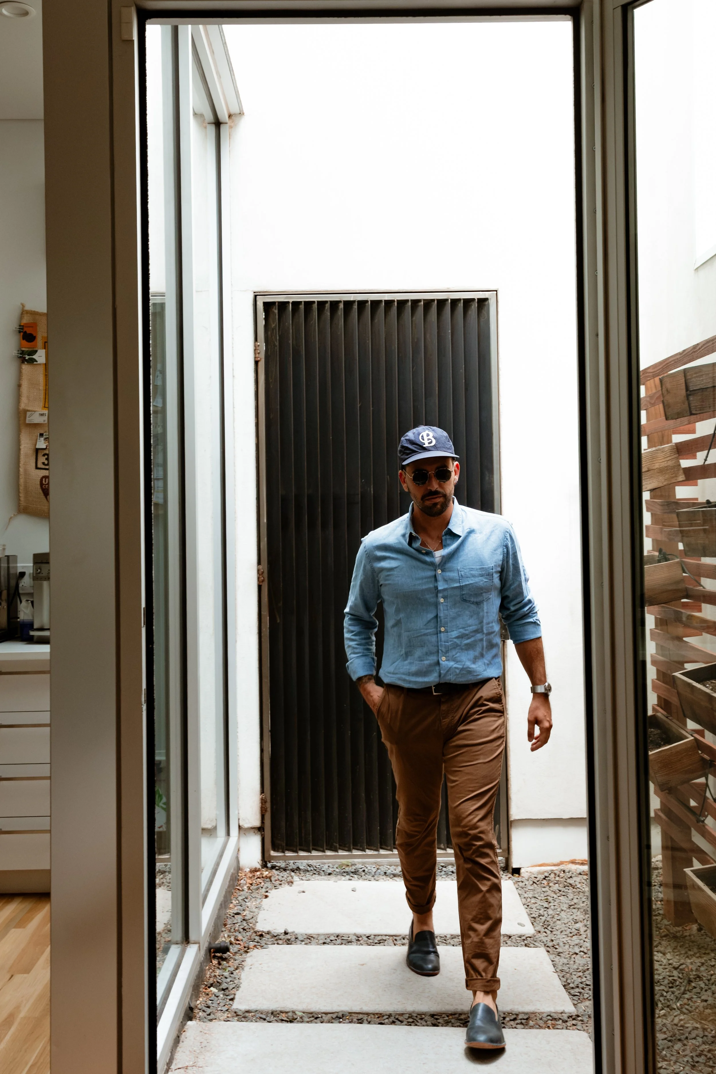Man walking out of a doorway into a small outdoor area. He is wearing sunglasses, a navy cap, a light blue button-up shirt, brown pants, and black shoes.