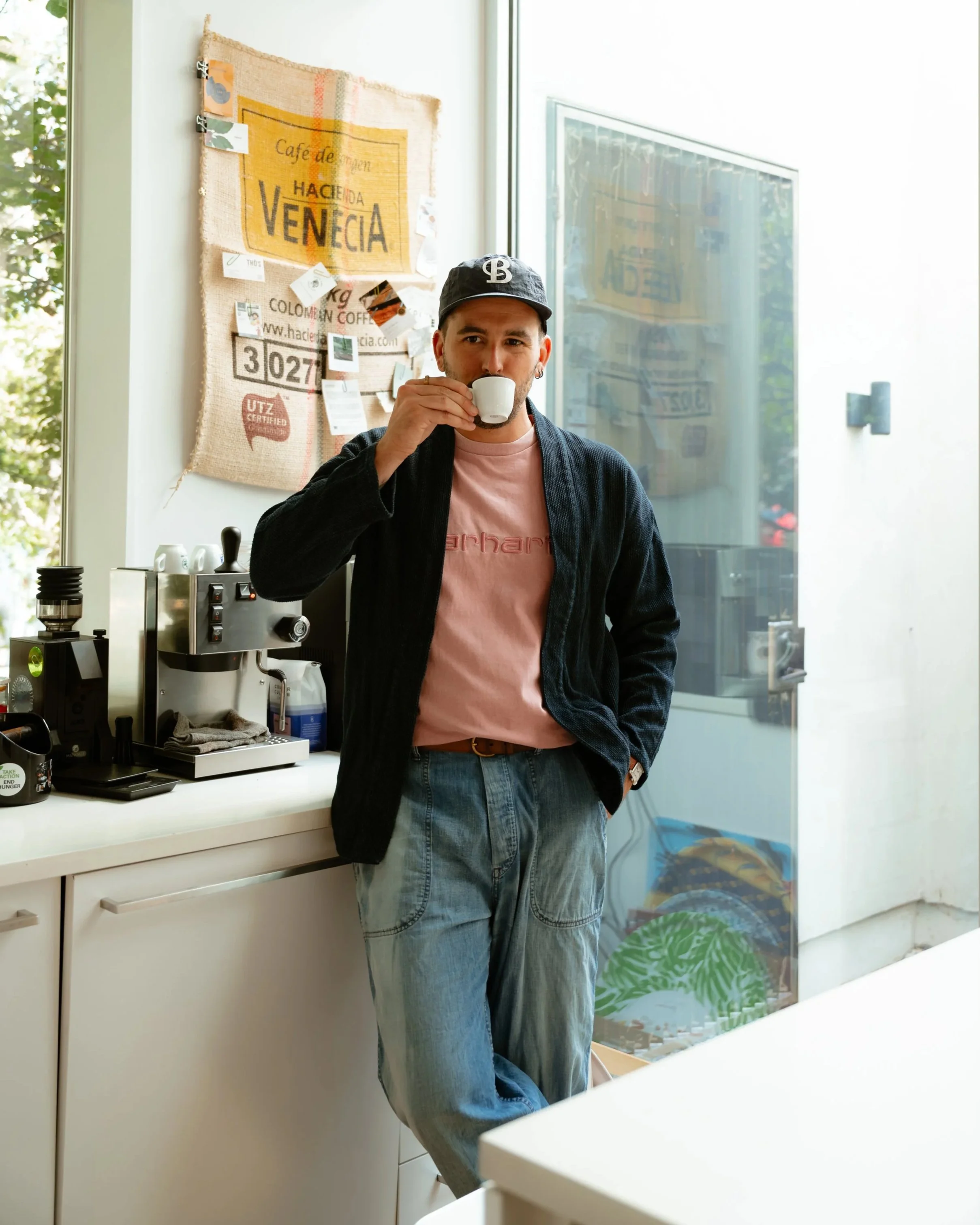 A man drinking espresso dressed in a timeless outfit with a dark blue jacket, light red t-shirt, loose-fitting jeans, in a modern kitchen with coffee equipment in the background.