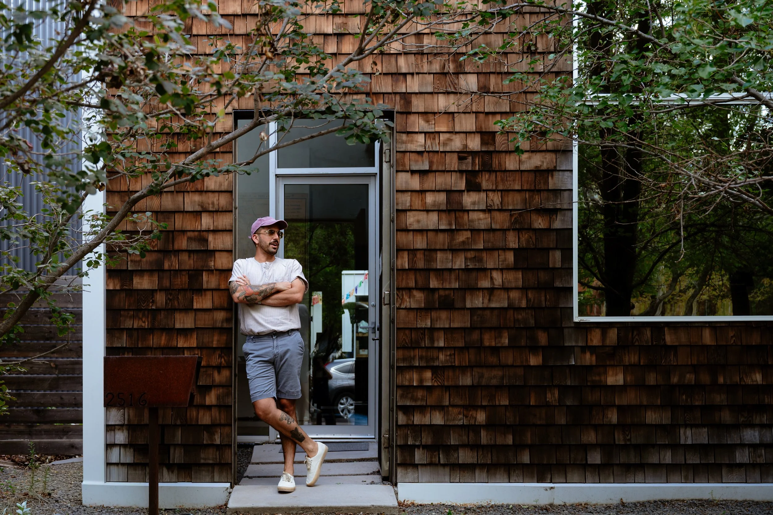 A man with tattoos wearing a pink cap and sunglasses, standing with arms crossed in front of a wooden house with large windows and a glass door, surrounded by trees.
