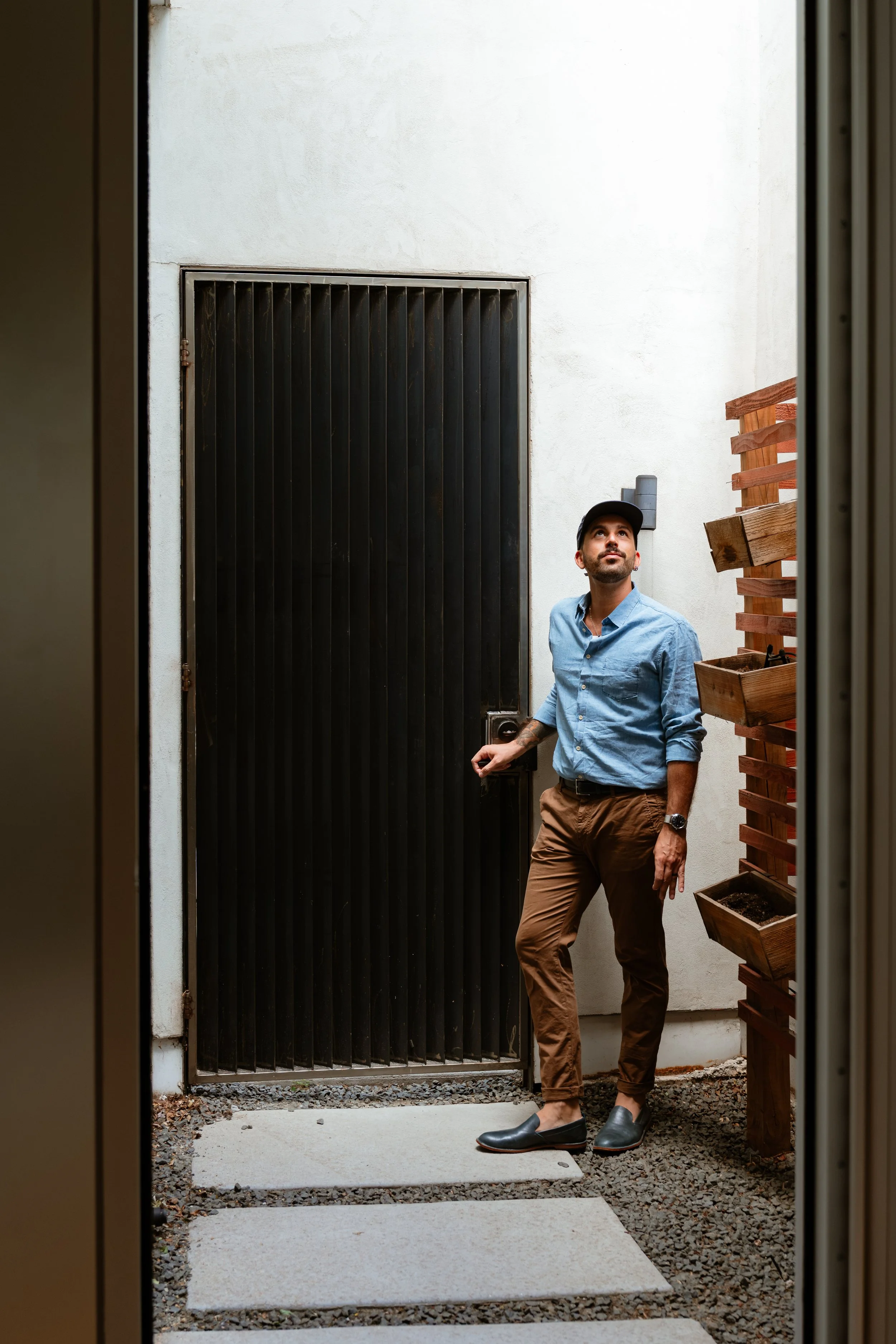 A man standing outside a door, holding a door handle, wearing a blue shirt, brown pants, black shoes, and a black cap, with a wooden lattice and wall behind him.