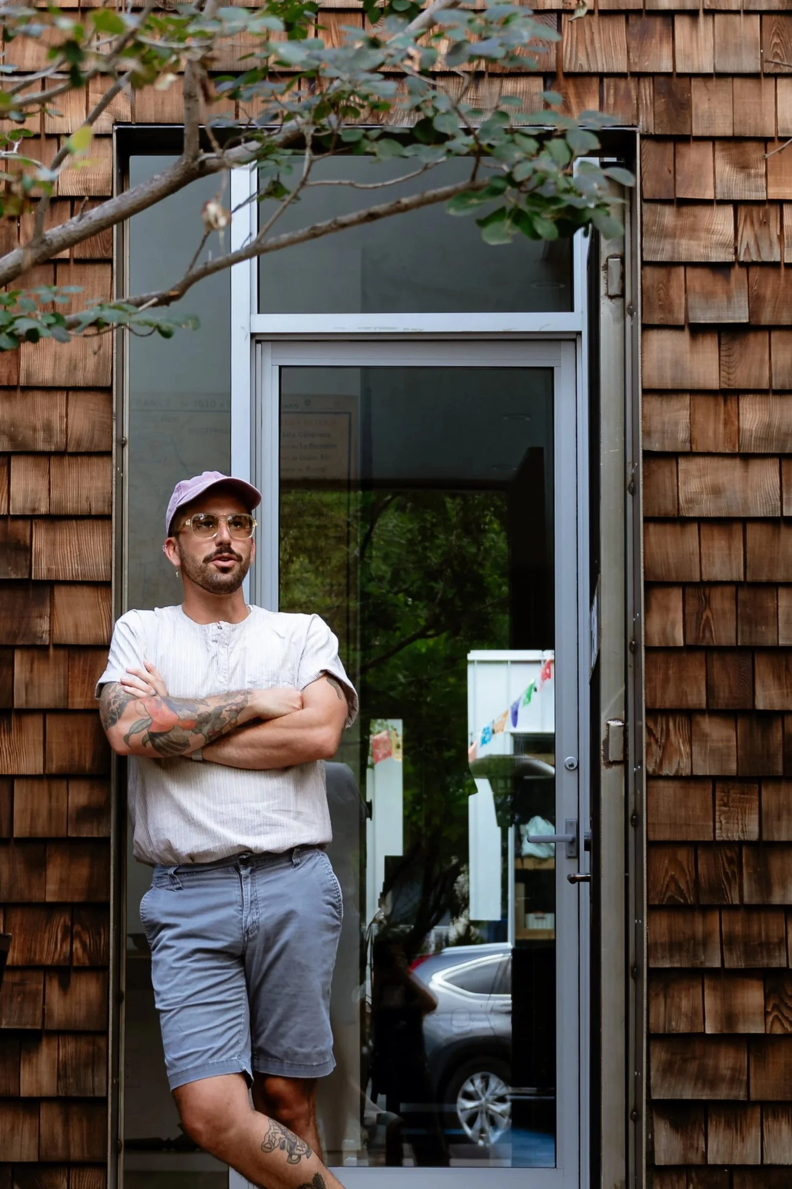 A man with glasses, a beard, and tattoos on his arm and leg, wearing a pink cap, standing with arms crossed outside a wooden building with a glass door, green leaves partially covering the top of the image, and a reflection of trees and a car in the glass door.