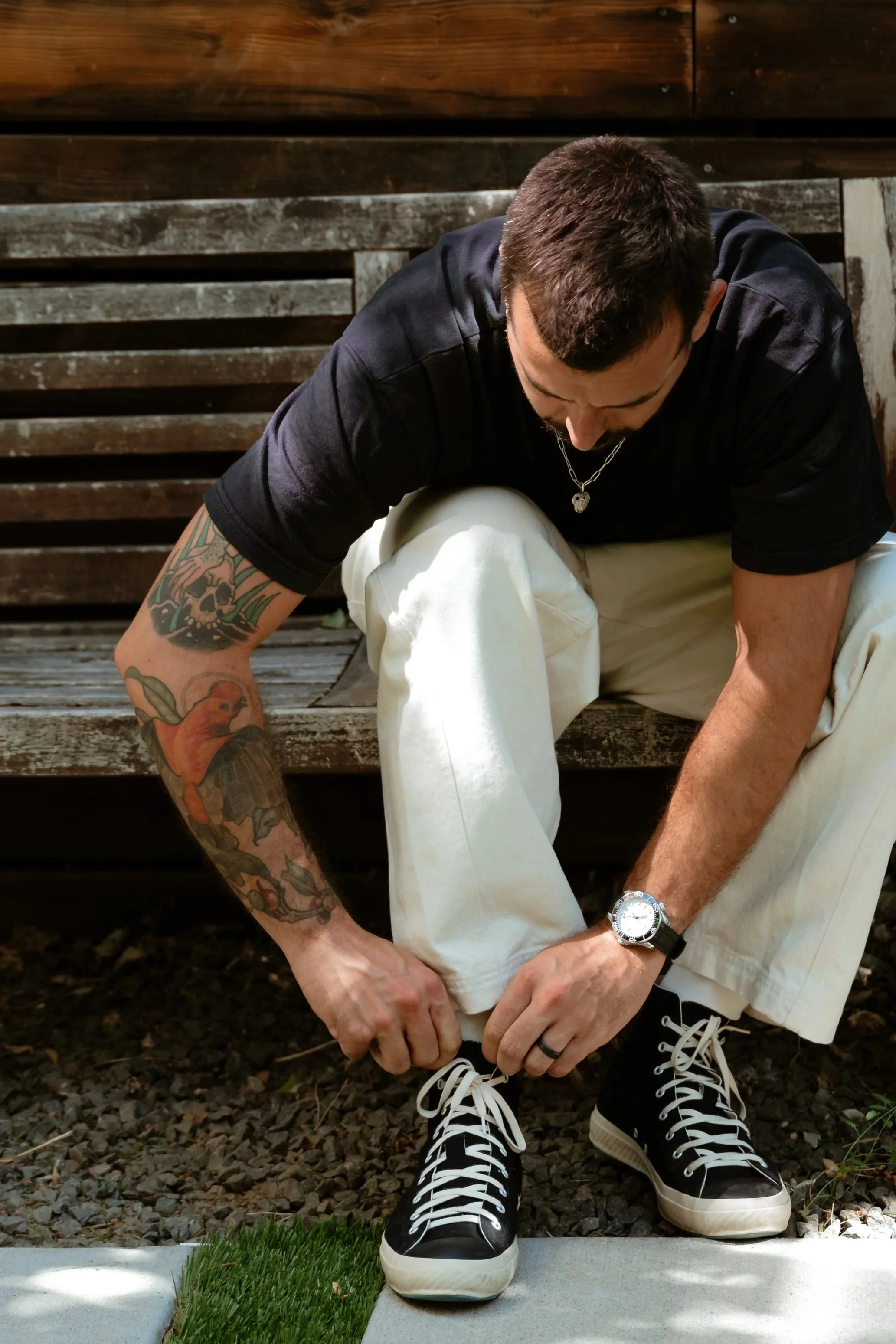 A man with tattoos on his arms is sitting on the ground tying his shoelaces. He is wearing a black t-shirt, light-colored pants, and black sneakers with white soles. He has a watch on his left wrist, a chain necklace, and short dark hair. The background shows a wooden fence or wall and the ground is covered in small rocks and dirt.