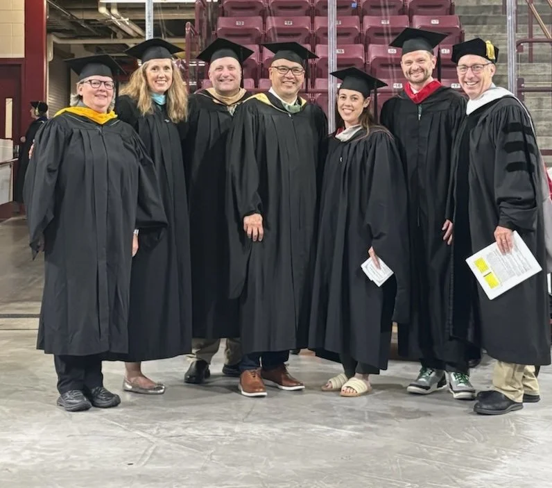 Group of seven graduates in caps and gowns standing in an indoor arena. Two women and five men, some holding diplomas, smiling for the photo.