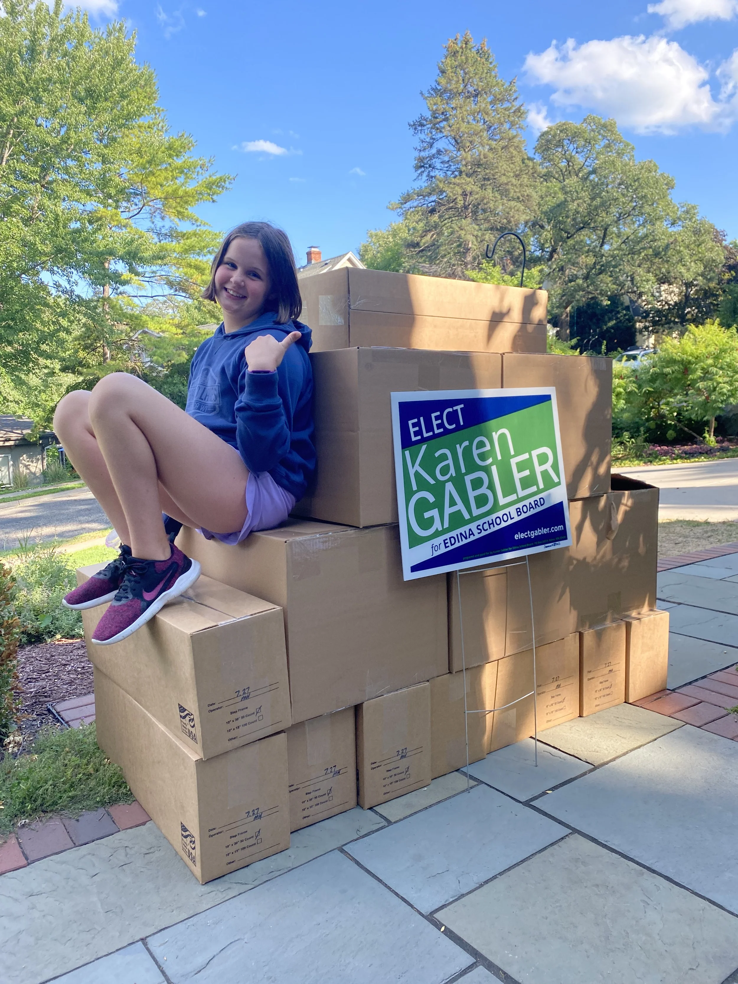 A smiling girl sitting on a stack of cardboard boxes outside on a sunny day with trees in the background. The boxes have a campaign sign for Karen Gabler for Edina School Board.
