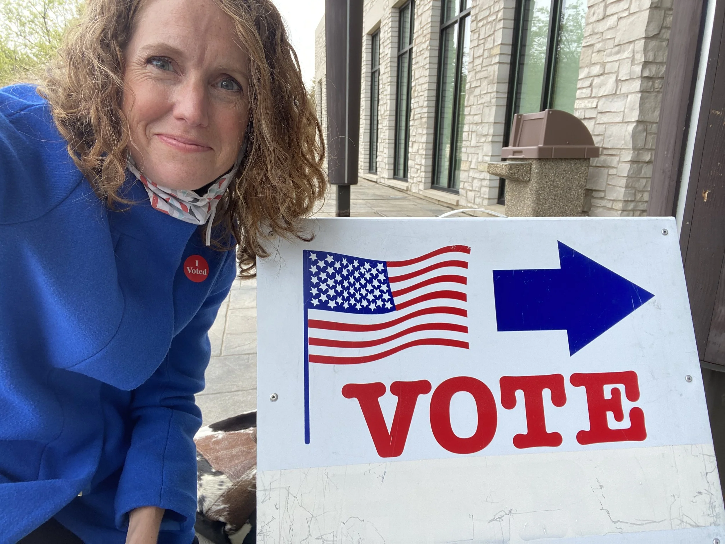 A woman with curly hair and a blue shirt wearing an 'I Voted' sticker takes a selfie beside a 'Vote' sign featuring the American flag and an arrow.