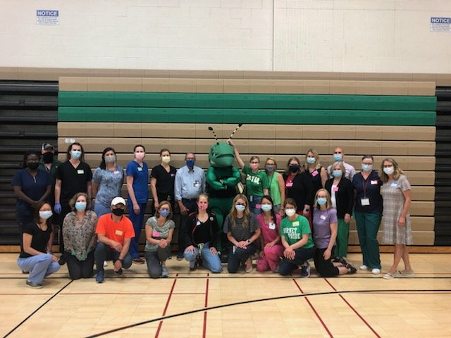 Group of people, including a person in a bug costume, gathered in a gymnasium with brown, green, and black bleachers in the background, all wearing masks.