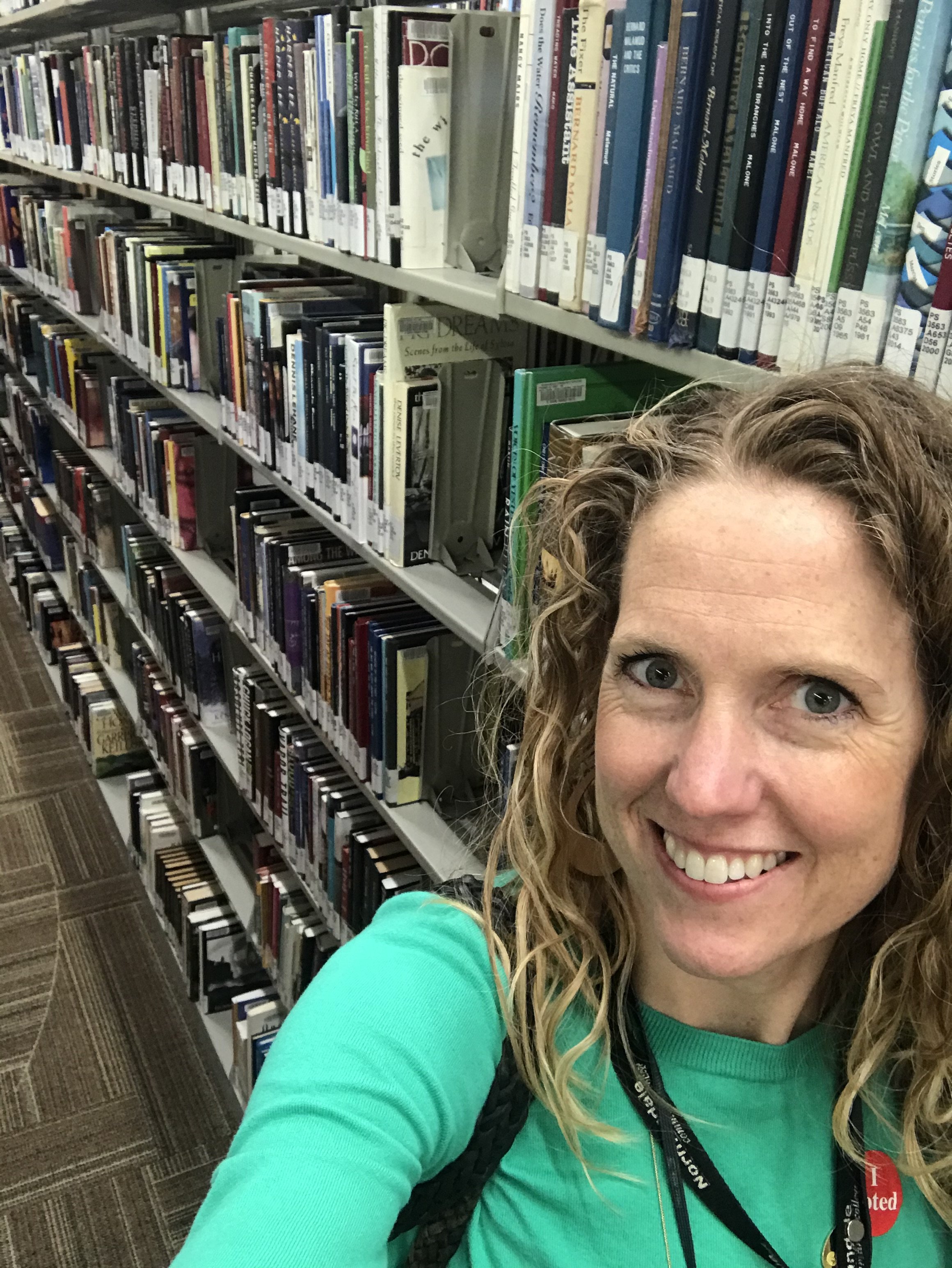 A smiling woman with curly blonde hair in a green shirt taking a selfie in a library aisle filled with shelves of books.