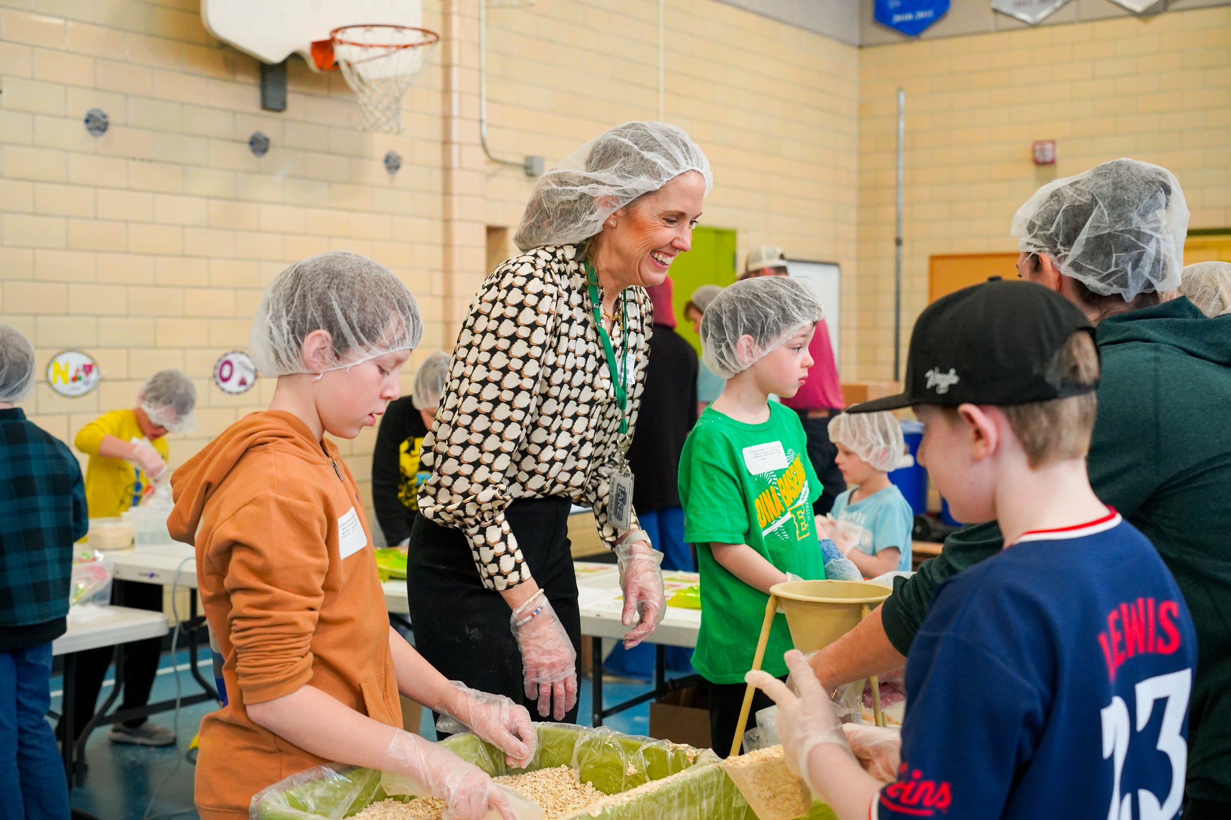A group of children and adults wearing hairnets and gloves are gathered around tables participating in a food packing activity in a gymnasium. They are filling bags with rice or similar food items.