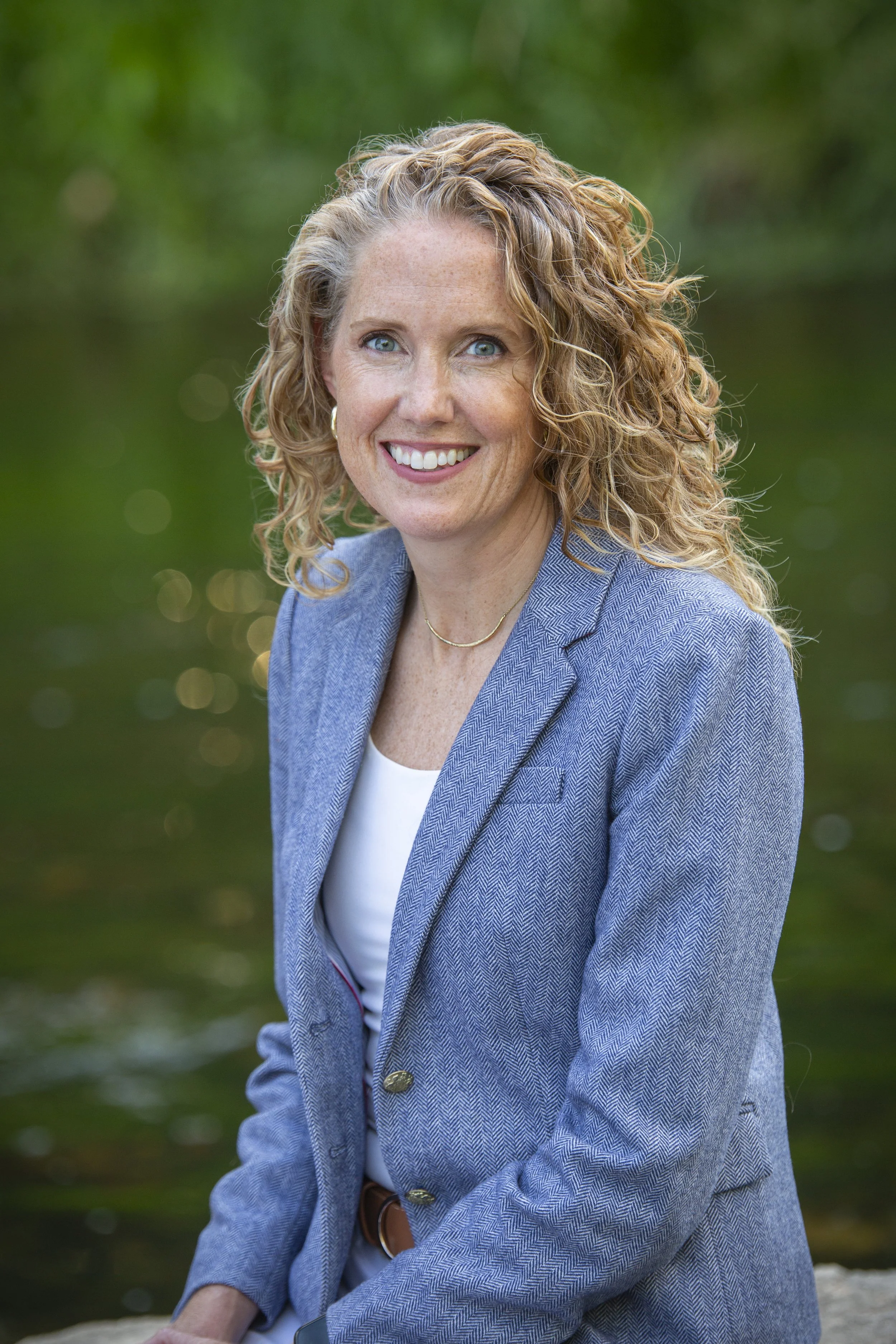 A woman with curly blonde hair and blue eyes smiling outdoors with a green, blurred background.