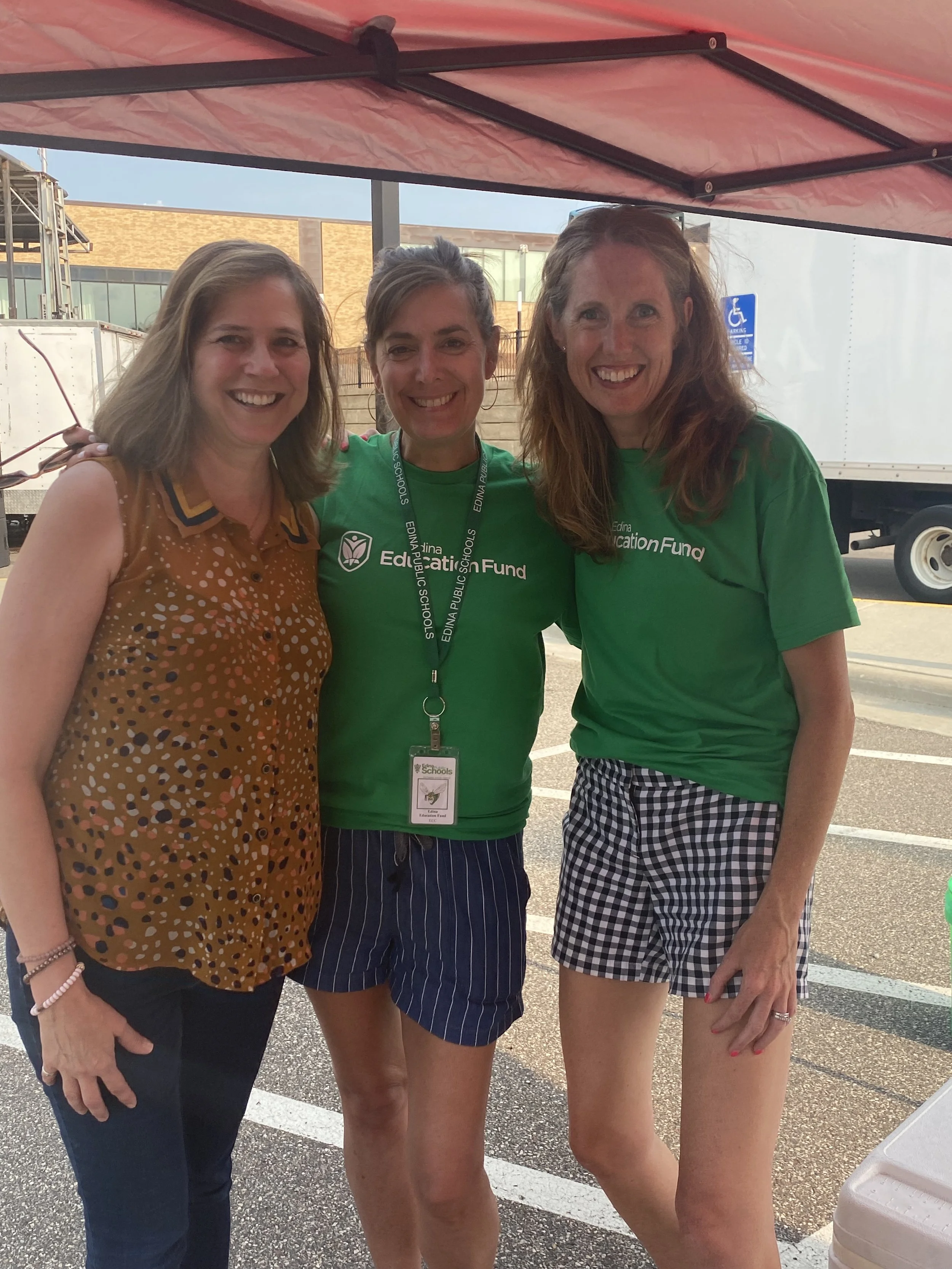 Three women standing close together outdoors in a parking lot under a canopy, smiling at the camera. All women have light skin and shoulder-length hair. The woman on the left is wearing a brown patterned sleeveless top, the woman in the middle is wearing a green t-shirt with a lanyard, and the woman on the right is also wearing a green t-shirt with plaid shorts.