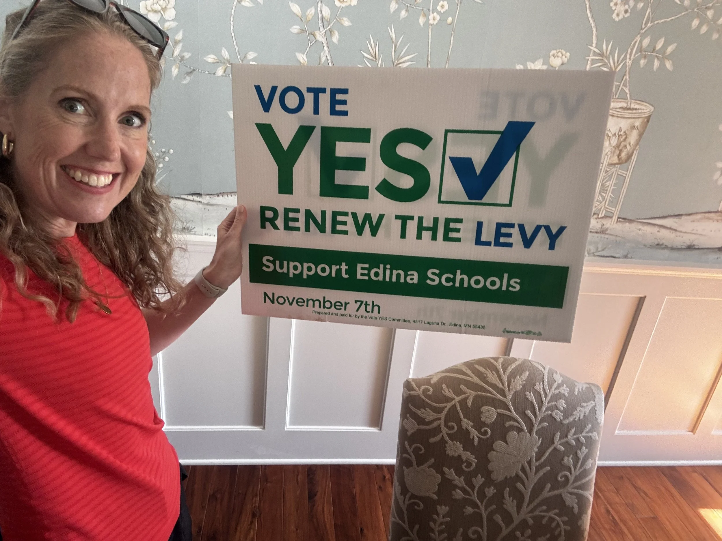 Woman smiling holding campaign sign that reads "Vote Yes Renew the Levy Support Edina Schools" in a room with patterned wallpaper and an upholstered chair.