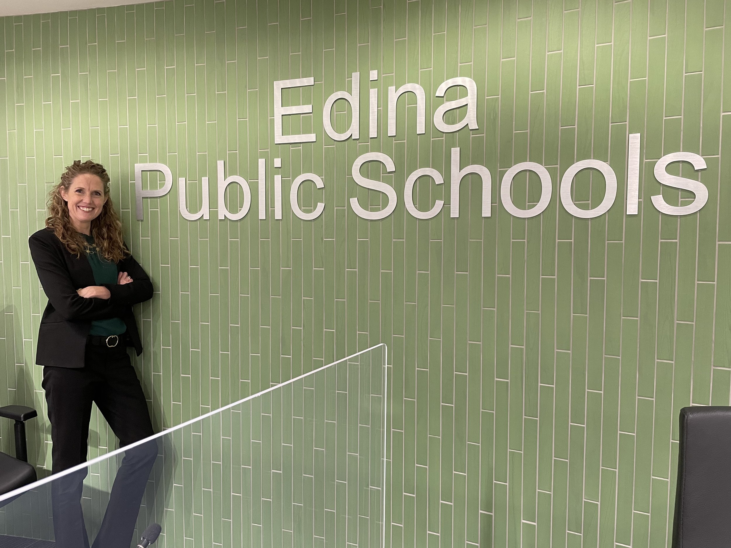 A woman with curly hair smiling and standing with crossed arms in front of a green tiled wall with large white letters reading 'Edina Public Schools'