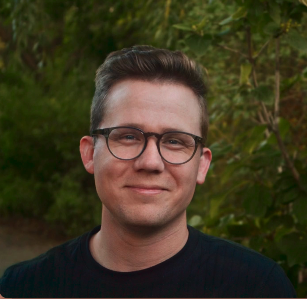 A man with short brown hair, glasses, and light skin, standing outdoors in front of green foliage.
