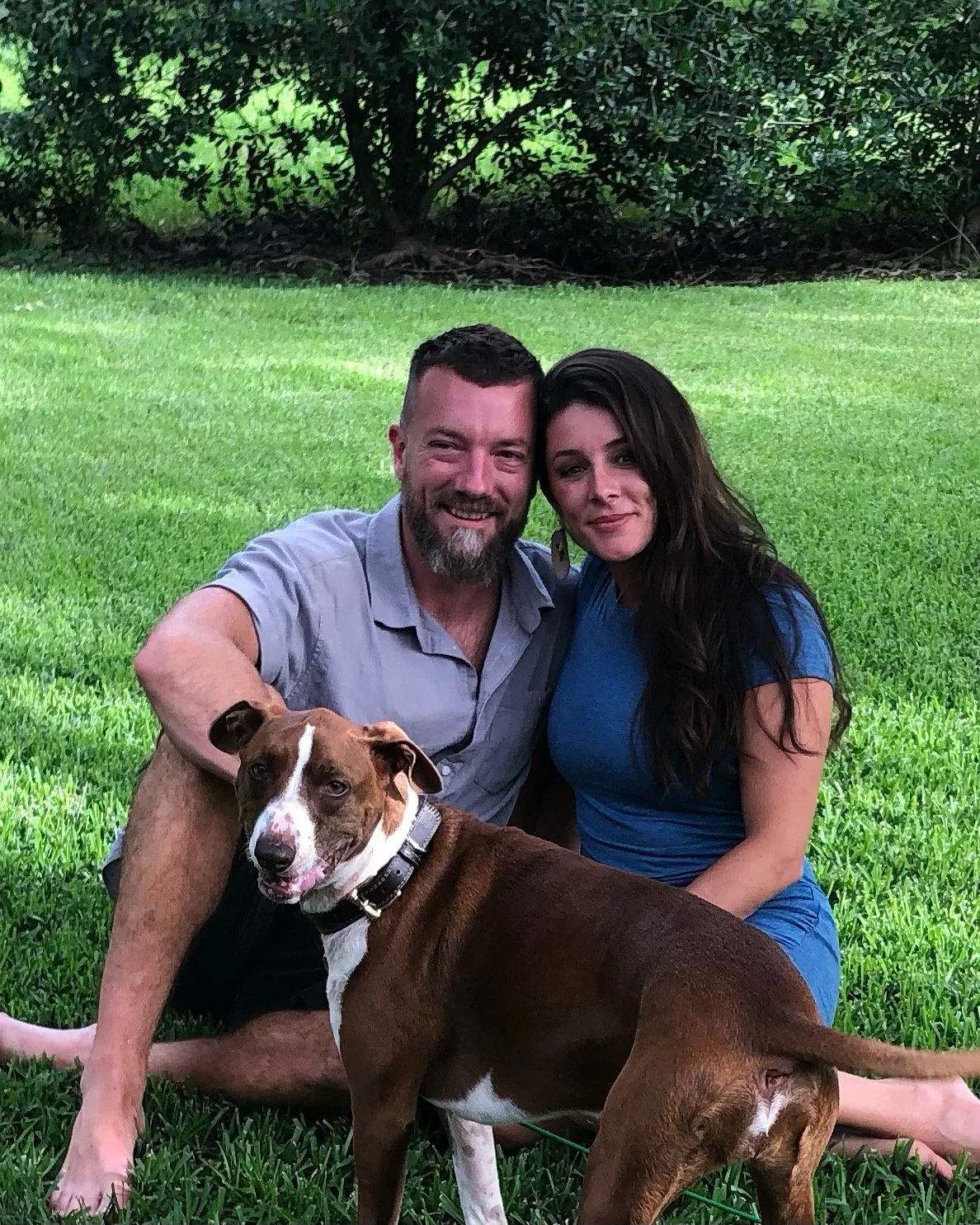 A man and woman sitting on grass with a brown and white dog in front of them, outdoors with trees in the background.