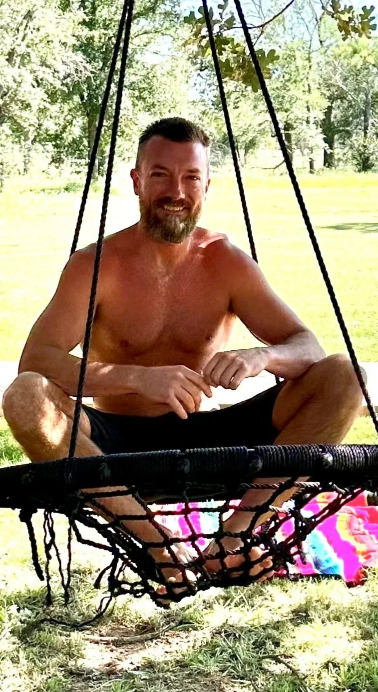 A shirtless man with a beard smiling while sitting cross-legged on a colorful beach towel under a tree, in front of a hanging hammock swing outdoors in a grassy area with trees in the background.