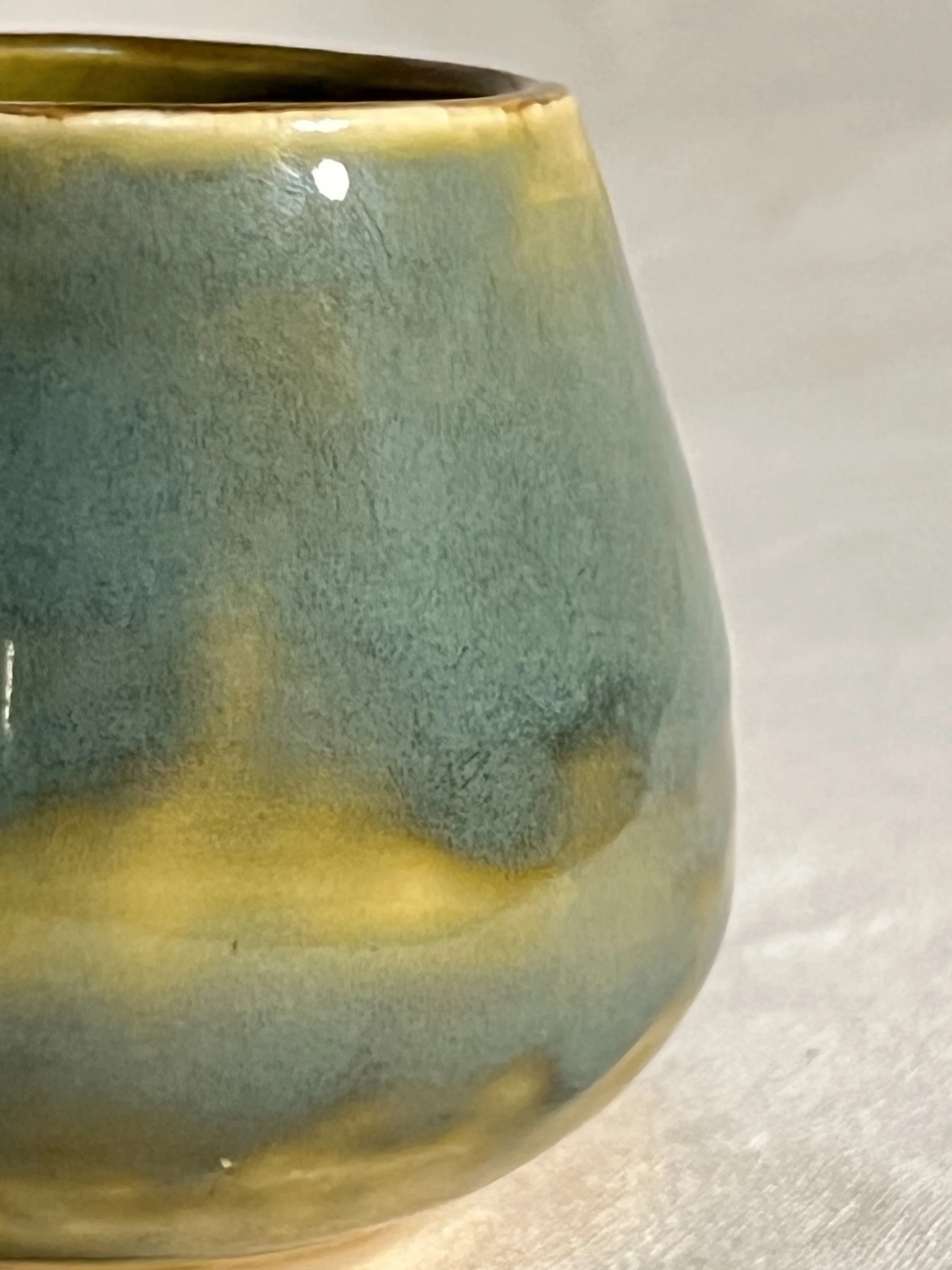 A closeup of a small round cup with glaze that varies between teal and yellow-green with a dripping pattern.