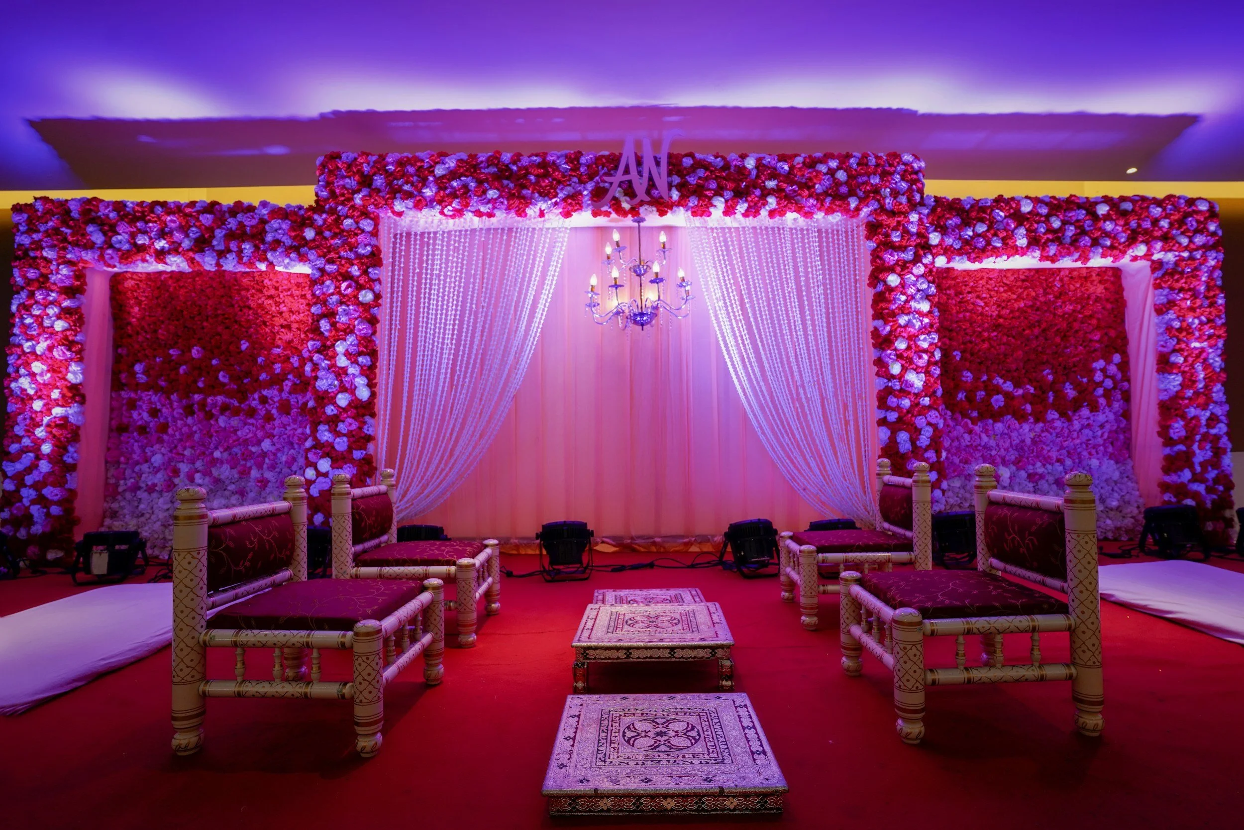 A decorated event stage with pink floral walls, a chandelier, draped pink fabric, and seating arrangements for a celebration.