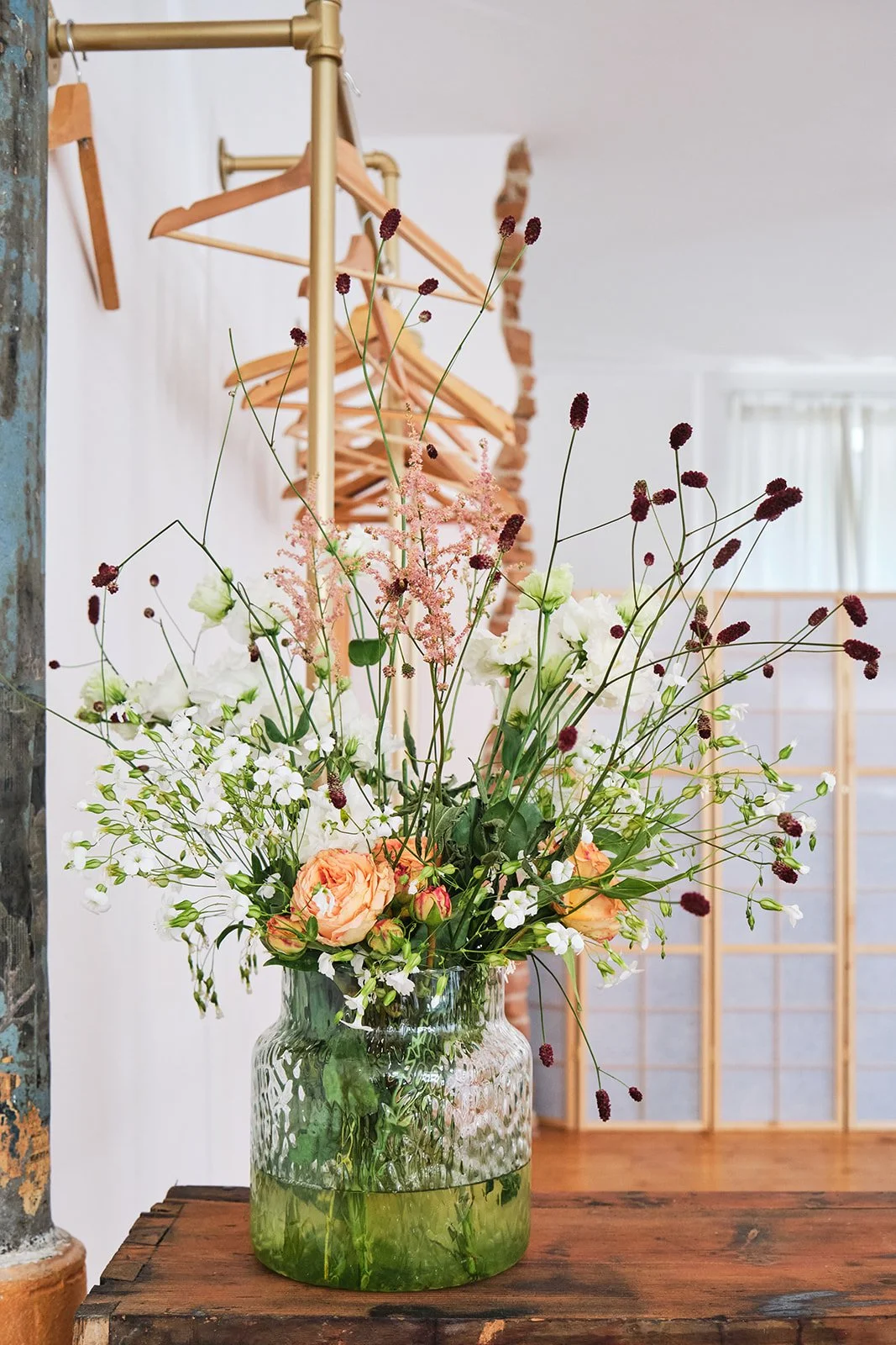Blumenstrauß mit weißen, peachfarbenen und dunkelroten Blumen in einer transparenten, grünlich schimmernden Vase auf einem Holztisch, im Hintergrund ein Kleiderständer und Raumteiler.