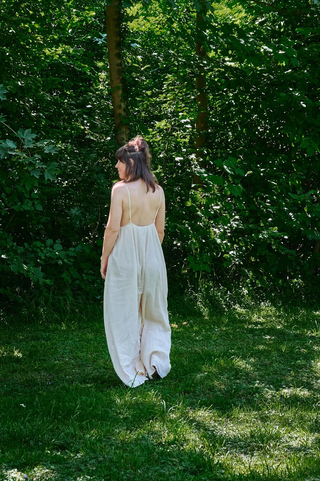 Eine Frau trägt einen weißen, lockeren, ärmellosen Schlafanzug und steht auf grünem Gras vor einer dichten, grünen Buschwand im Freien bei Sonnenlicht.