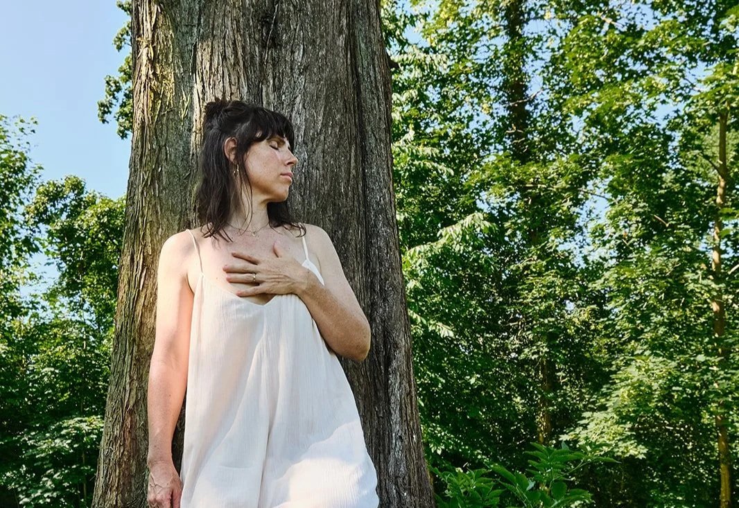 Eine Frau in einem weißen Kleid lehnt entspannt an einer Baumstamm in einem grünen Wald, mit geschlossenen Augen.