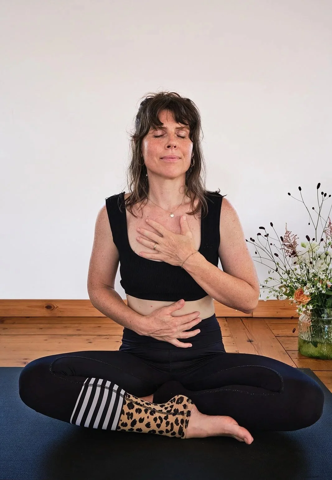 Eine Frau sitzt entspannt im Lotussitz auf einer Yogamatte, hält ihre hand auf die Brust und den Bauch, während sie bei einer Meditation mit geschlossenem Blick in einem Raum mit Holzfußboden und Blumen im Hintergrund sitzt.