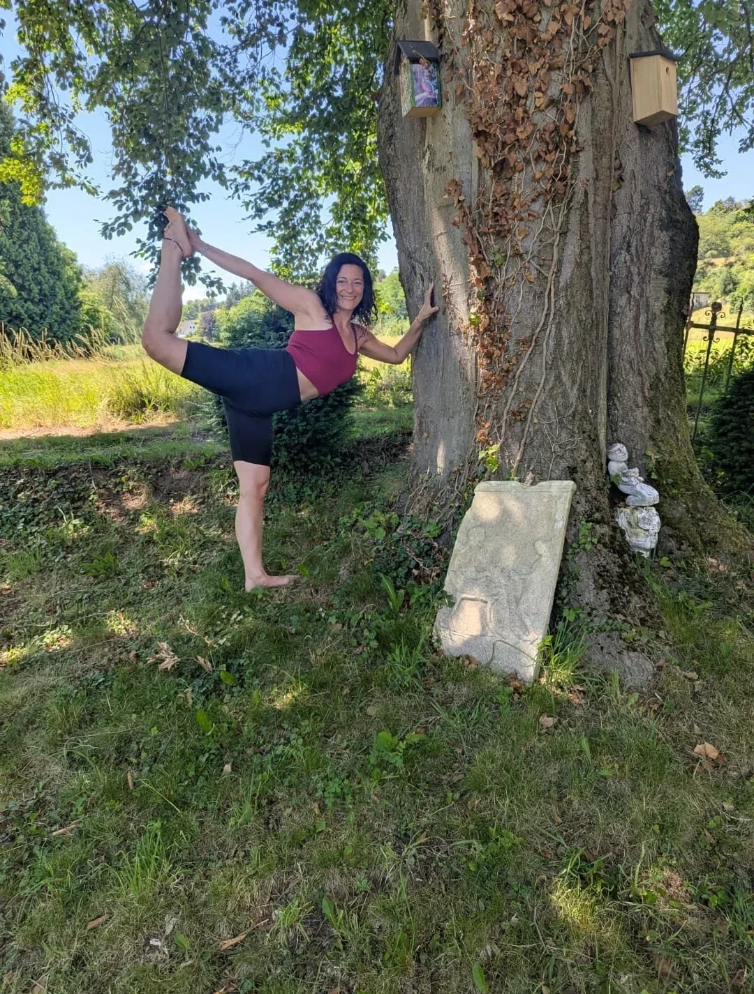 Frau macht Yoga im Grünen neben einem großen Baum mit Vogelnistkasten und Skulpturen.