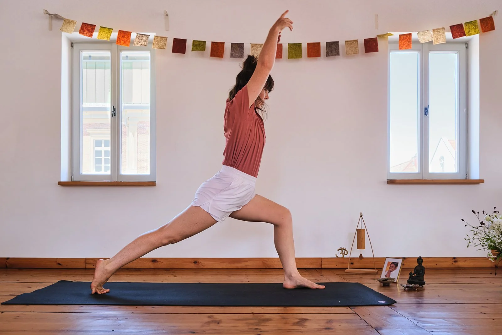 Frau macht Yoga in einem Raum mit Holzboden, zwei Fenster und bunten Wimpeln an der Wand.
