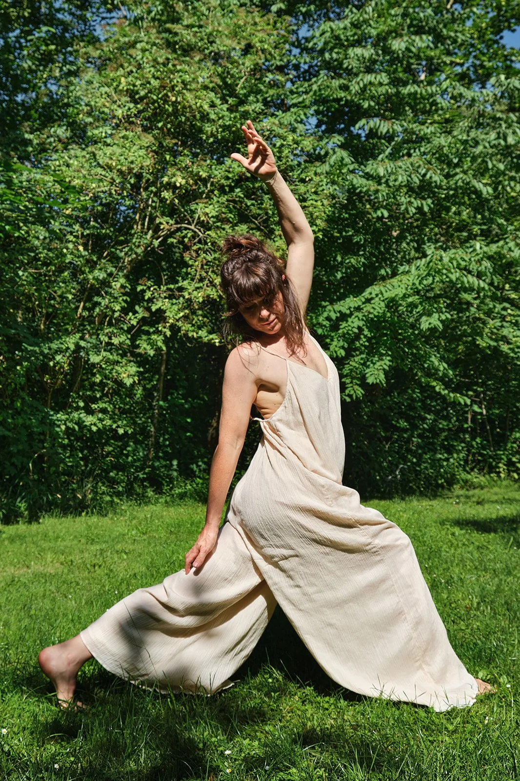 Eine Frau trägt ein langes, beige Kleid und pose in einem grünen Garten mit Bäumen im Hintergrund, während sie einen Kunst-Tanzhaltung einnimmt.