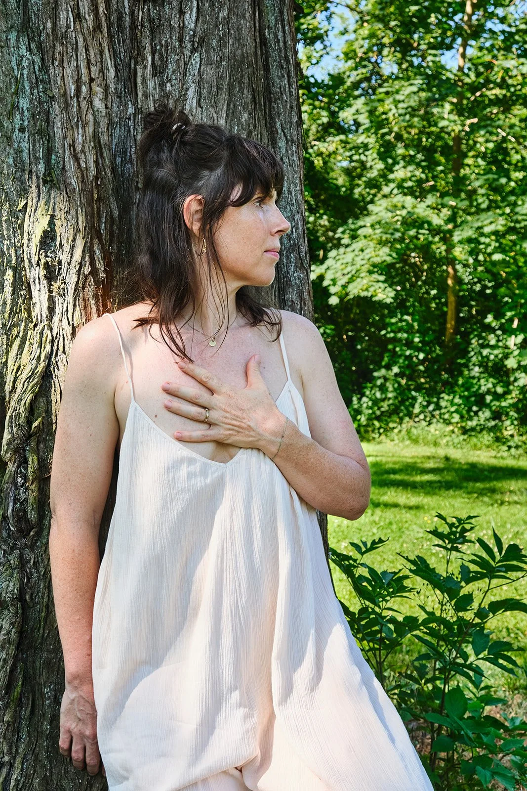 Eine Frau in einem weißen Kleid lehnt an einem Baum im Grünen, hält die Hand auf die Brust und schaut nach rechts.