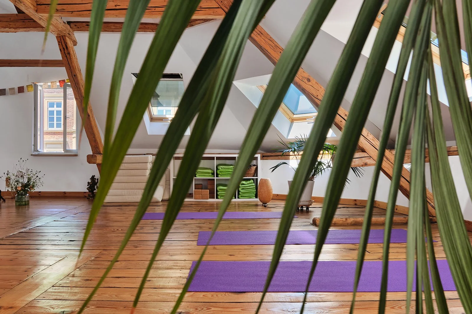 Ein heller, gemütlicher Yoga-Raum im Dachgeschoss mit Holzfußboden, großen Fenstern und Skydachfenstern, eingerichtet mit Yogamatten, Pflanzen, einem Regal mit grünen Handtüchern und Dekorationen.