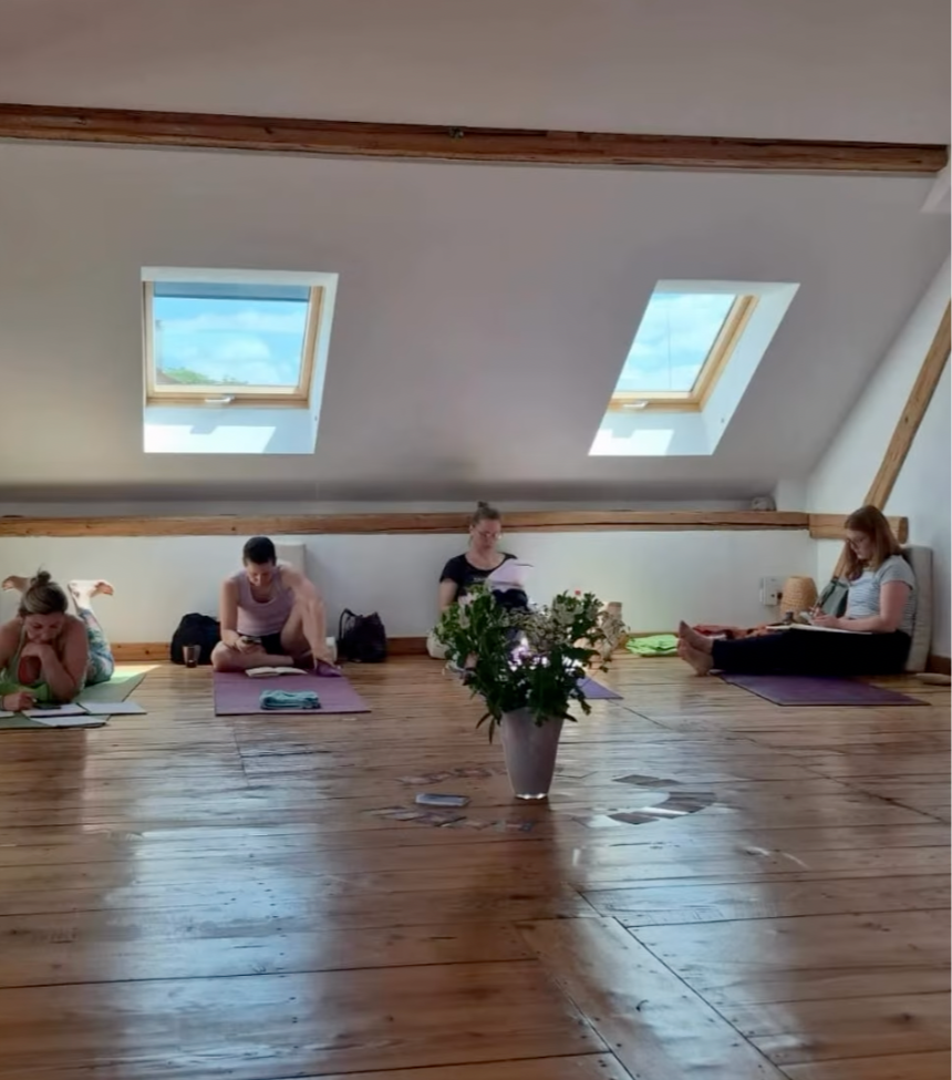 Vier Frauen sitzen auf Yogamatten in einem Raum mit Holz Böden und Dachfenstern, eine Vase mit Blumen steht in der Mitte des Raumes.
