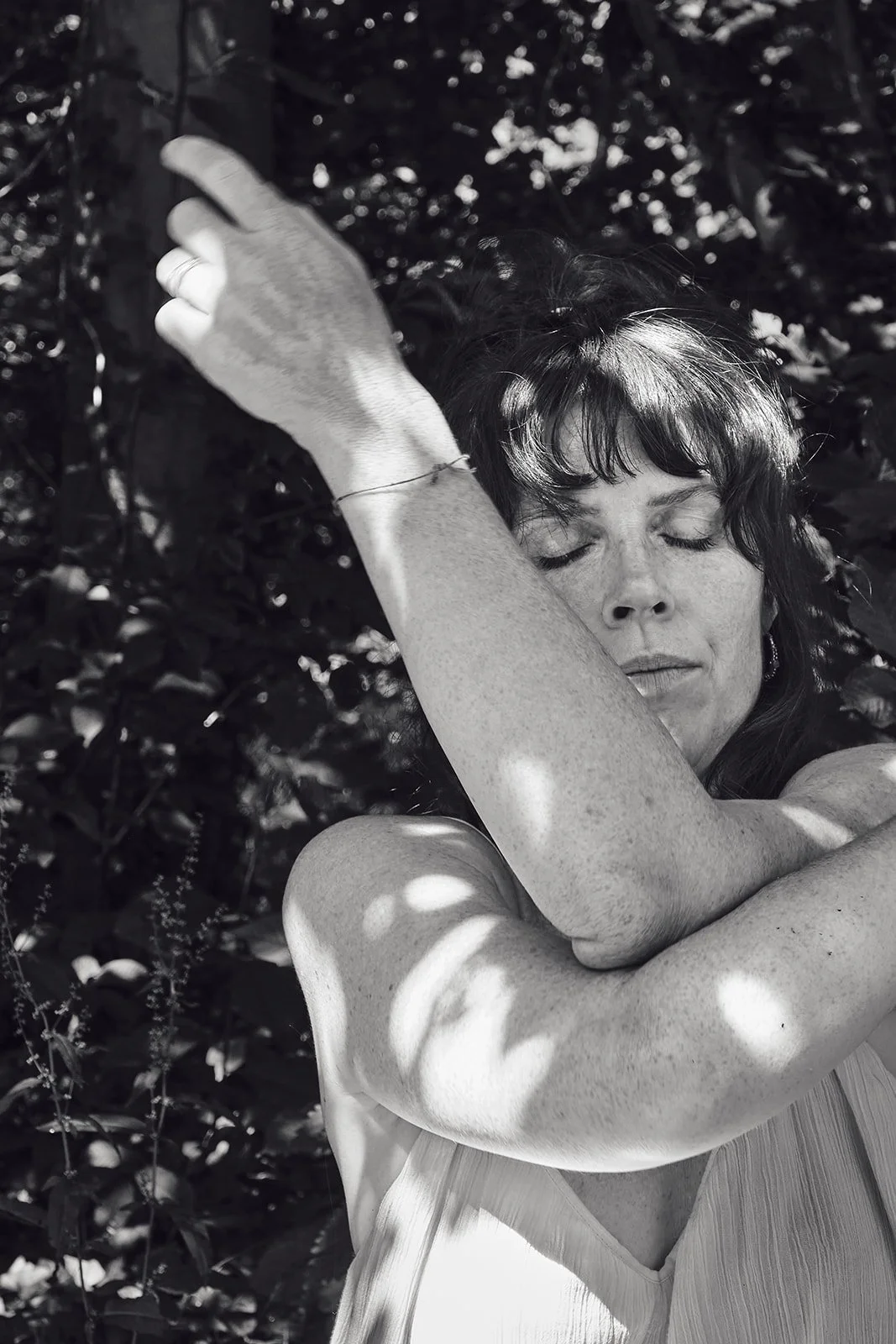 Schwarz-weiß-Foto einer Frau mit geschlossenen Augen, die sich an einem Baum oder Ast festhält, im Hintergrund dichtes Blattwerk.