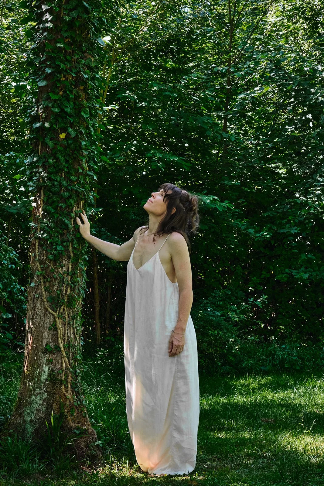 Eine Frau in einem weißen Kleid steht im Grünen und berührt einen Baum, im Hintergrund sind viele Bäume und Blätter.